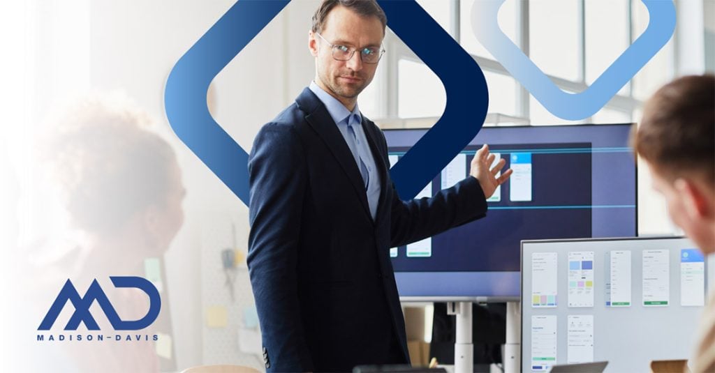 A man in a business suit and glasses speaks to two others, gesturing to a large computer monitor
