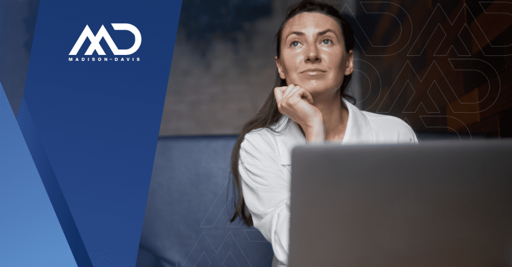 Viewed from behind a laptop screen, a woman in corporate attire pauses to think.