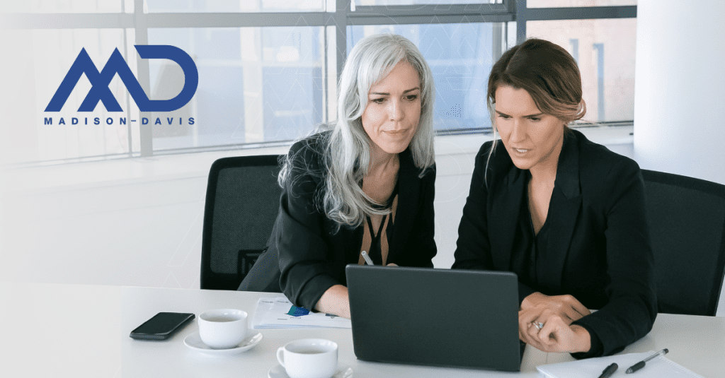 Two women in corporate attire discuss over a laptop at a table with coffee, notebooks, pens, and papers.