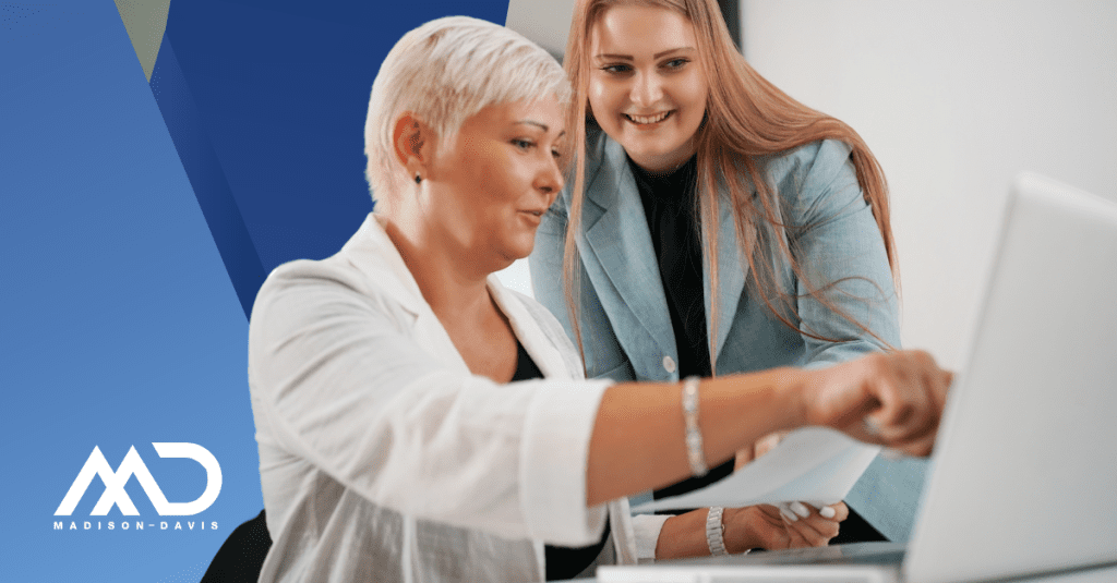 A veteran worker and younger employee in corporate attire discuss the content on a paper and laptop screen