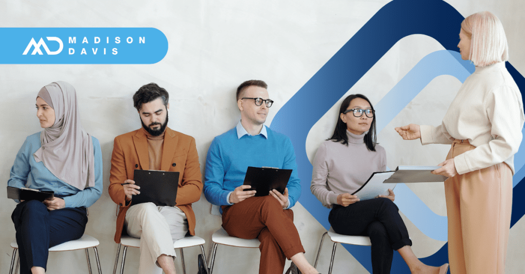 Four applicants in corporate attire sit in a row holding clipboards. One recruitment professional holding a folder stands addressing them.