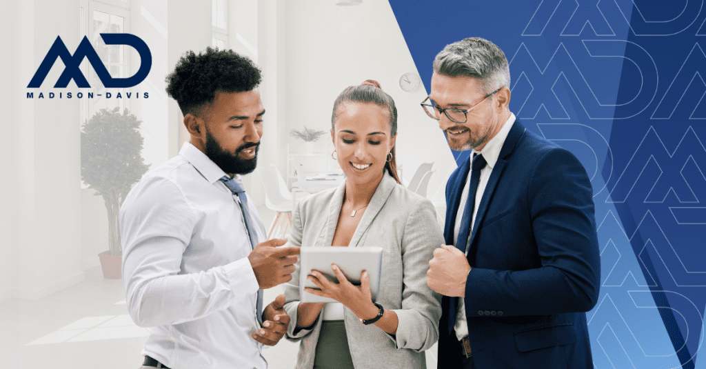 Three people in corporate attire with ties and blazers discuss over a tablet