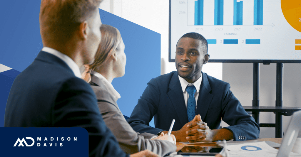 Man in corporate attire sits at a conference table speaking to two coworkers, the table and flat display screen showing various graphs