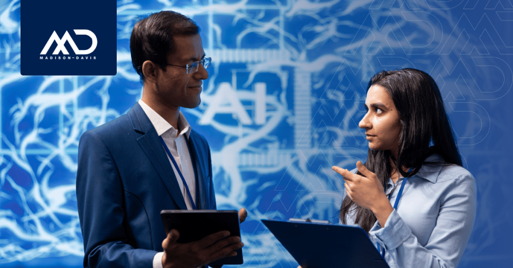 Man in corporate attire holding a tablet speaks to a woman in corporate attire holding a clipboard