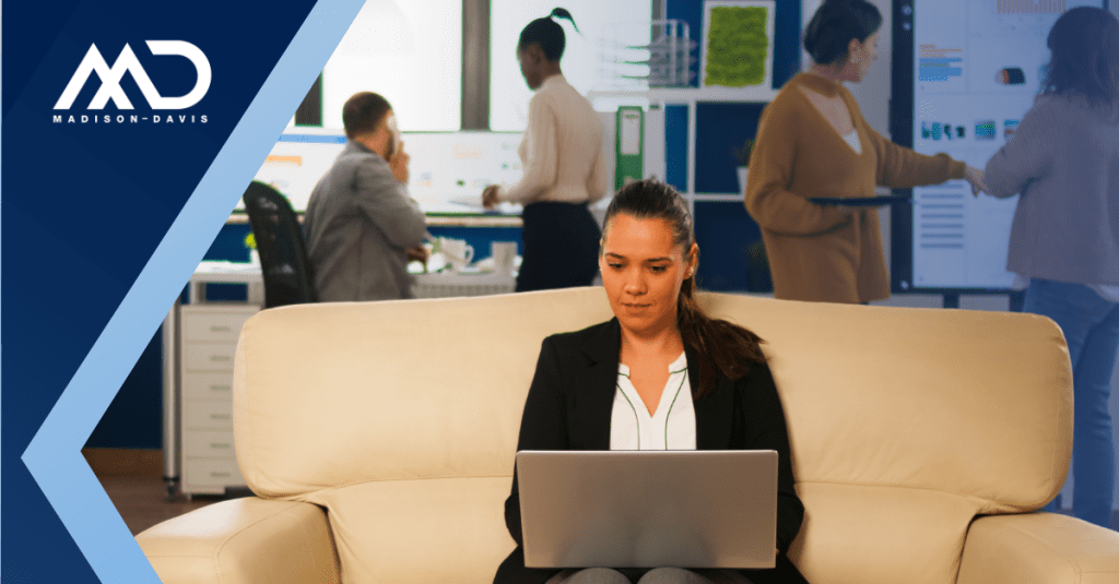 A woman in corporate attire sits on a couch with a laptop on her lap, her back to a busy office environment